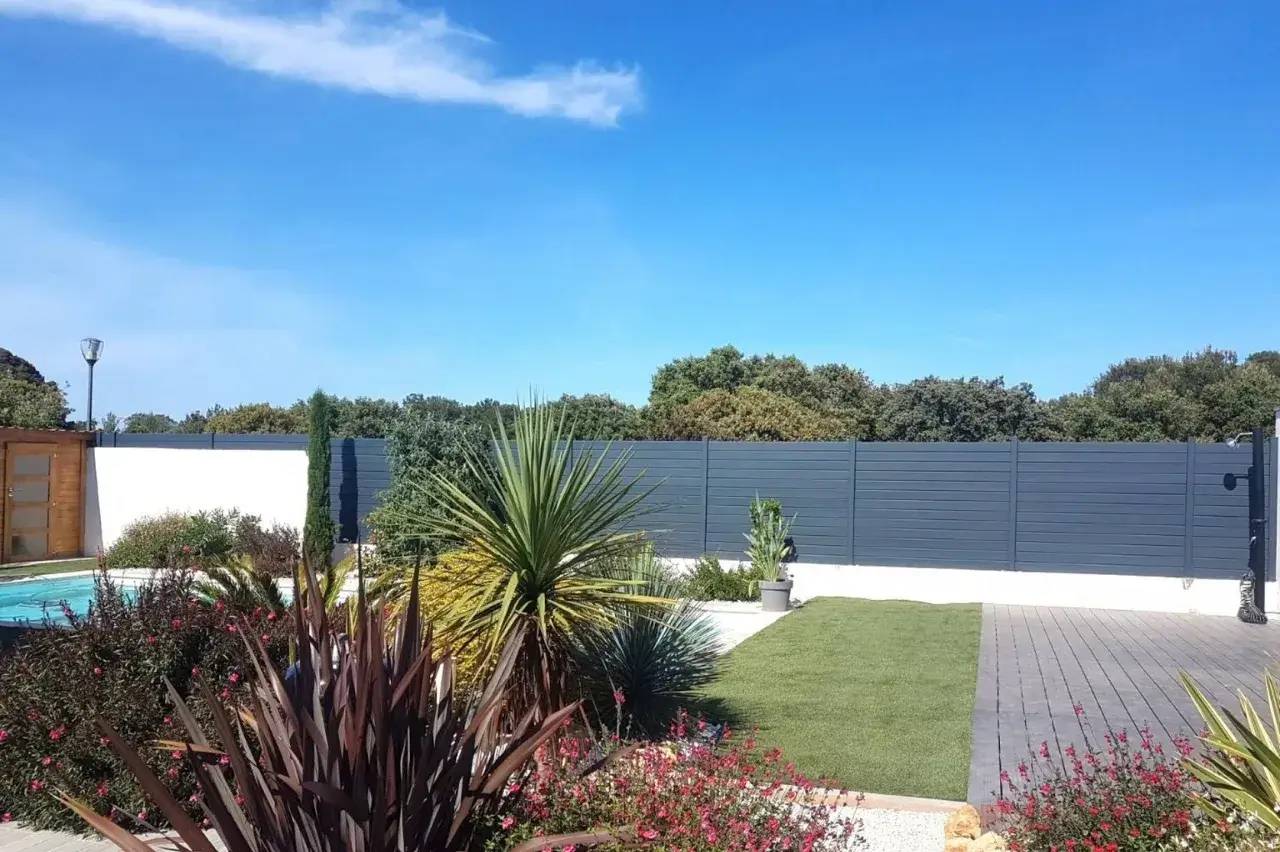 Une cour extérieure élégante avec des plantes tropicales, une pelouse soignée et une clôture moderne sous un ciel bleu vif.
