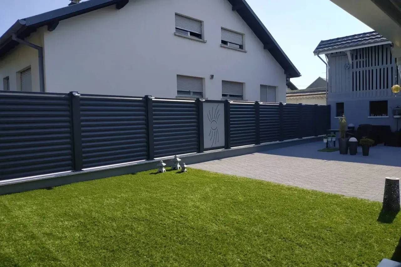 Une maison moderne entourée d'une clôture métallique sombre se dresse près d'une pelouse verte bien entretenue et d'une cour pavée.