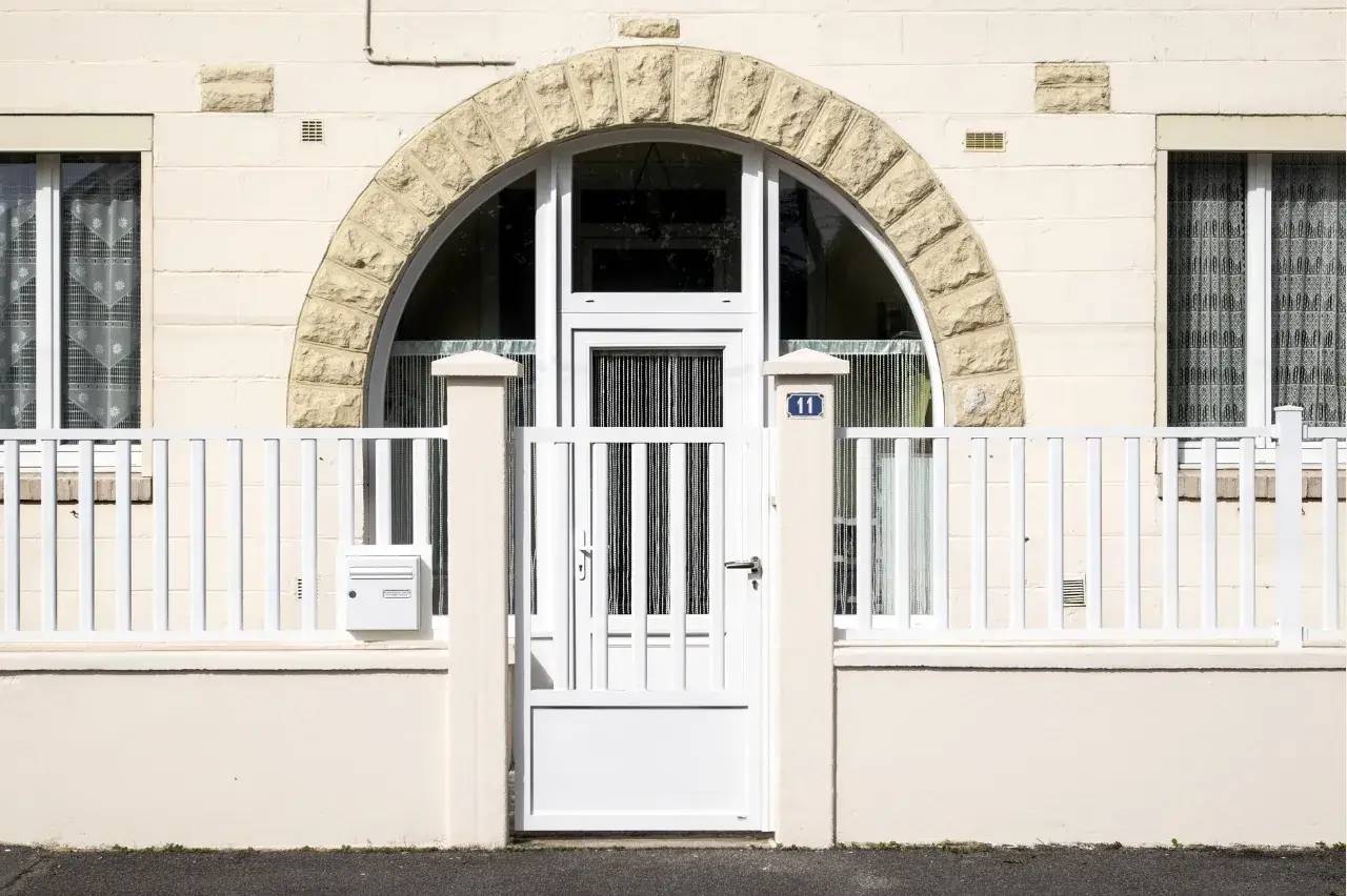Façade élégante en pierre claire avec une porte blanche arquée encadrée de grilles blanches et de fenêtres en verre.