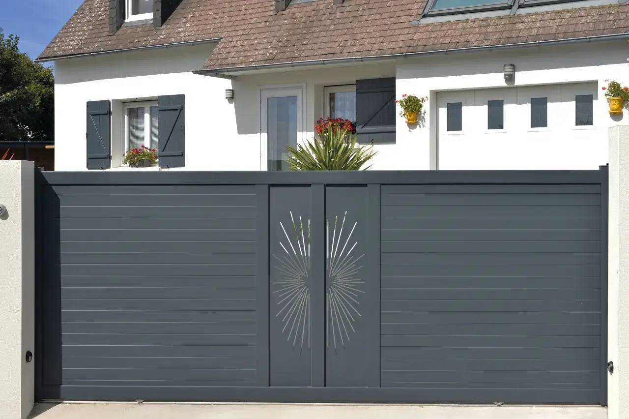 Un portail moderne gris anthracite avec un motif central ajouré, devant une maison blanche aux volets foncés.