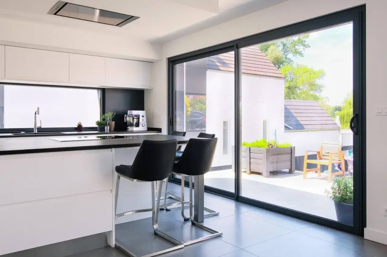 Cuisine moderne avec îlot central, comptoir noir et grandes baies vitrées donnant sur une terrasse ensoleillée.