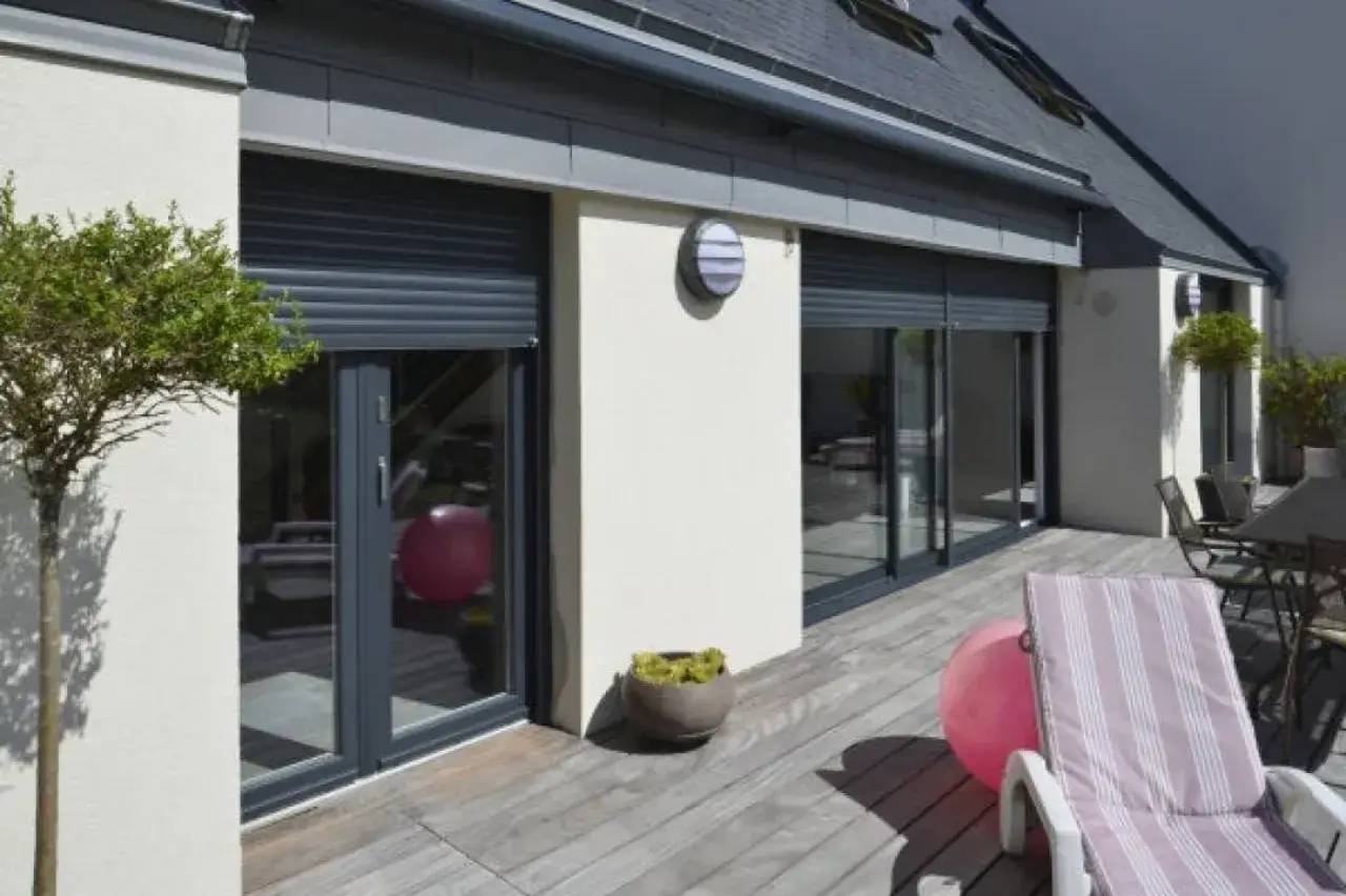 Une terrasse en bois ensoleillée avec des chaises longues rayées, entourée de grandes baies vitrées et de plantes en pot.
