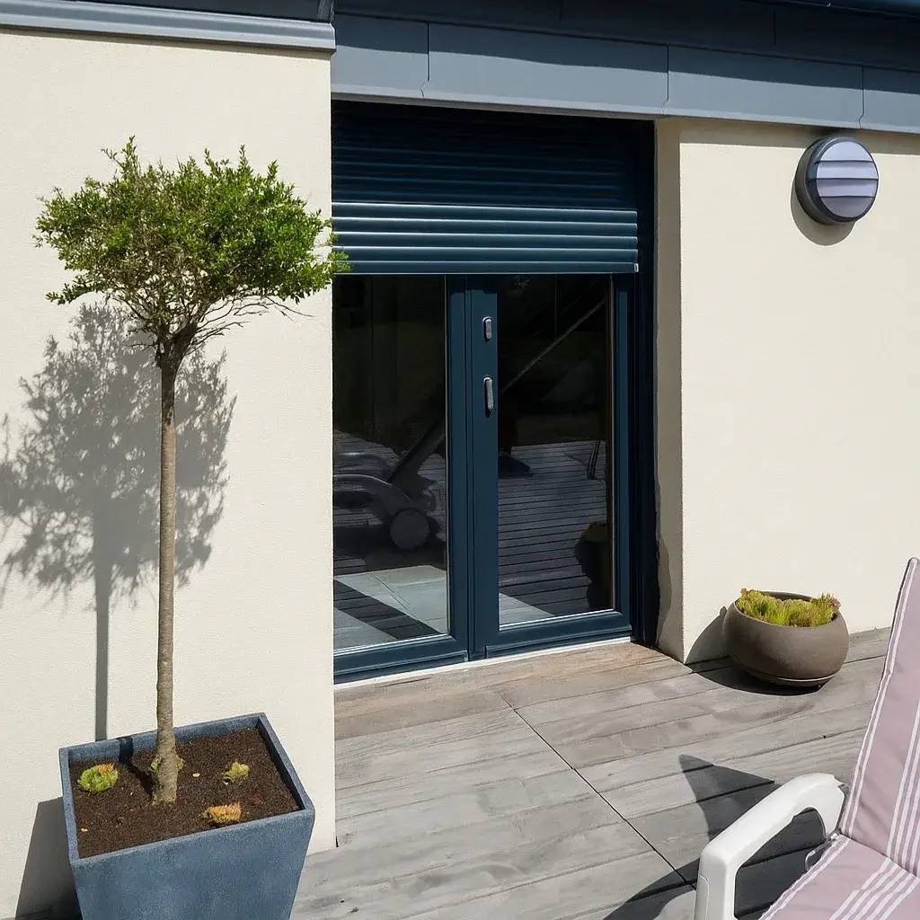 Une terrasse ensoleillée avec un petit arbre en pot à gauche, une porte vitrée bleue, et une chaise longue partiellement visible à droite