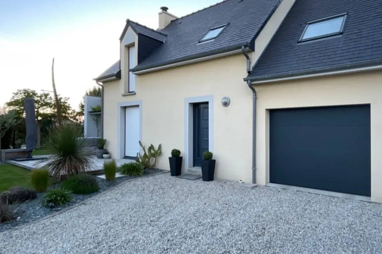 Une maison moderne aux murs beige avec un toit en ardoise, un garage et une entrée sur un jardin paysager en gravier.